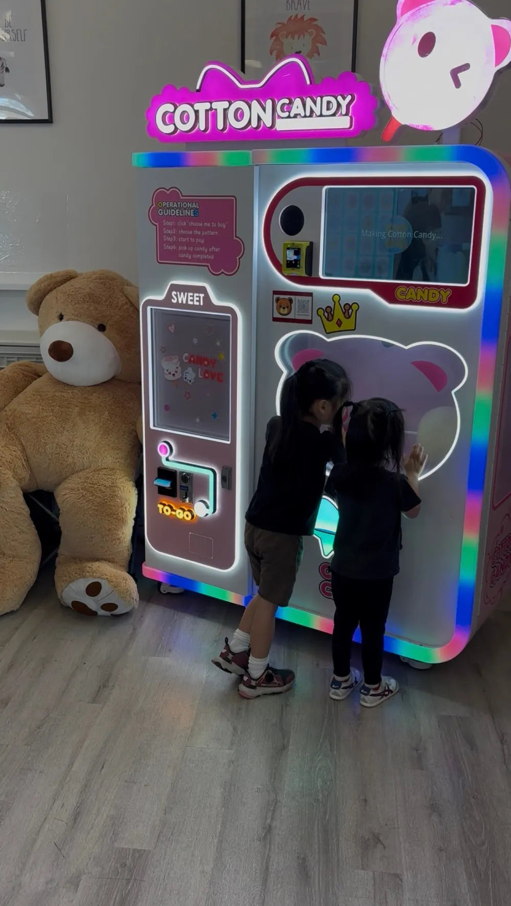 The LED cotton candy machine lit up, with two children at the screen and a giant teddy bear beside it